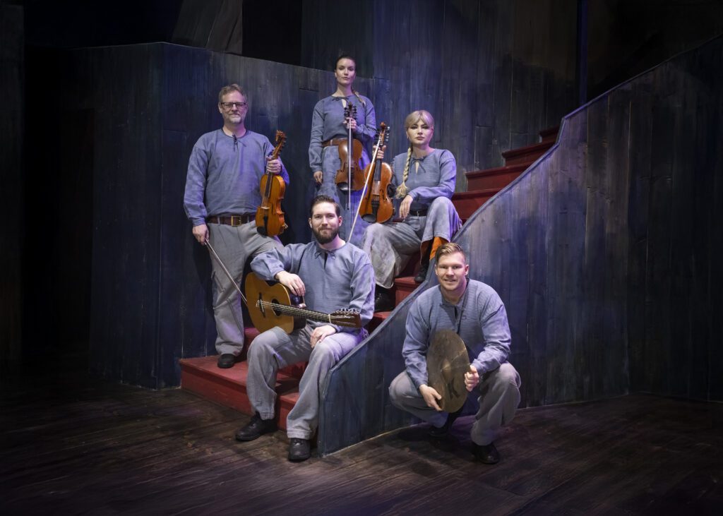 Foto med fem musiker i blå kläder. Tre stycken håller i varsin fiol, en annan har en gitarr och den femte håller i en del av ett trumset.