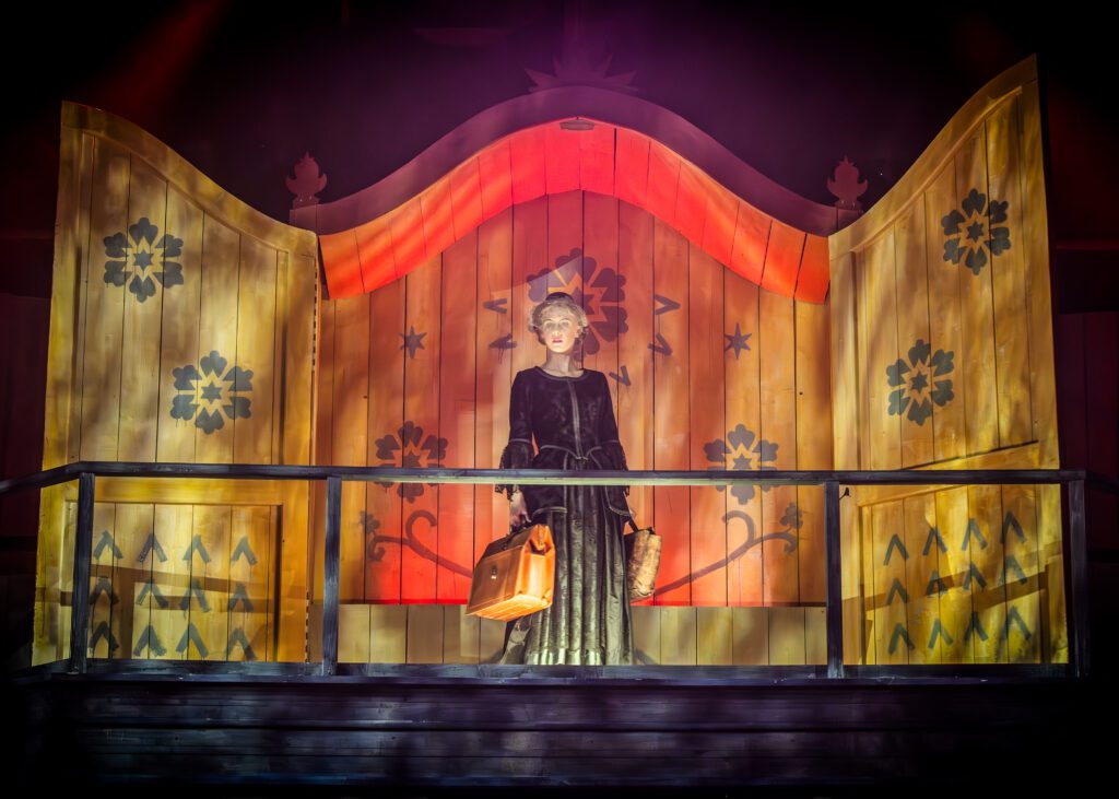 Foto från Kejsarn av Portugallien. Klara Gulla, spelad av Edith Nilsson, i svarta kläder med en resväska i handen. I bakgrunden finns klara, starka färger.